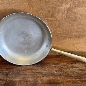 May include: A silver-colored frying pan with a long, gold-toned handle. The pan is round with a flat cooking surface and a slightly raised edge. The handle has a loop at the end for hanging. The pan is set against a wooden background.