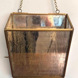 May include: A vintage, hexagonal-shaped hanging planter with a brass frame and a mirrored interior. The planter is suspended by a delicate brass chain. The glass has a distressed, aged appearance, adding to its antique charm. The planter is empty.
