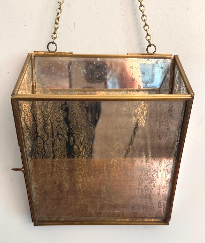 May include: A vintage, hexagonal-shaped hanging planter with a brass frame and a mirrored interior. The planter is suspended by a delicate brass chain. The glass has a distressed, aged appearance, adding to its antique charm. The planter is empty.