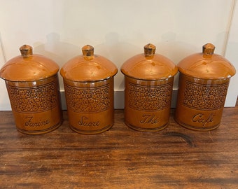 May include: Four matching ceramic canisters with lids. The canisters are a warm brown color with a floral pattern and the words "Farine", "Sucre", "Thé", and "Cafe" in black script. The lids have a small knob.