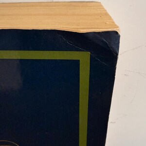 Może przedstawiać: Zbliżenie rogu książki. Książka ma ciemnoniebieską okładkę z zieloną obw&oacute;dką i złotym wzorem. Strony są w jasnobeżowym kolorze. Książka jest ustawiona na białym tle.