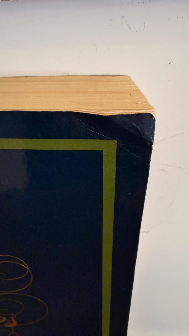 Może przedstawiać: Zbliżenie rogu książki. Książka ma ciemnoniebieską okładkę z zieloną obw&oacute;dką i złotym wzorem. Strony są w jasnobeżowym kolorze. Książka jest ustawiona na białym tle.