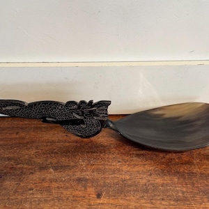 May include: A dark-colored serving spoon with a dragon-shaped handle. The spoon's bowl is oval-shaped and has a gradient of colors. The handle is intricately carved with a dragon design, adding a decorative element to the utensil.
