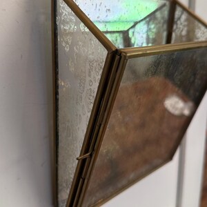 May include: A geometric, hanging planter with a mirrored glass design and a brass-colored metal frame. The planter is suspended by a delicate chain. The glass has a mottled, antique appearance, adding a vintage aesthetic. The planter is empty.
