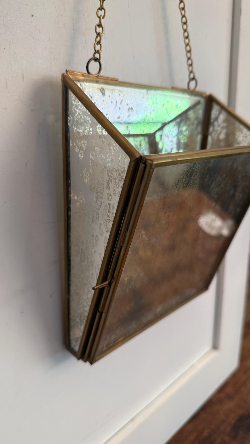 May include: A geometric, hanging planter with a mirrored glass design and a brass-colored metal frame. The planter is suspended by a delicate chain. The glass has a mottled, antique appearance, adding a vintage aesthetic. The planter is empty.