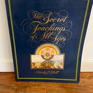 Może przedstawiać: Książka w twardej oprawie zatytułowana "The Secret Teachings of All Ages" autorstwa Manly'ego P. Halla. Książka ma ciemnoniebieską okładkę ze złotymi literami i kolorową ilustracją. Książka leży na drewnianej powierzchni.