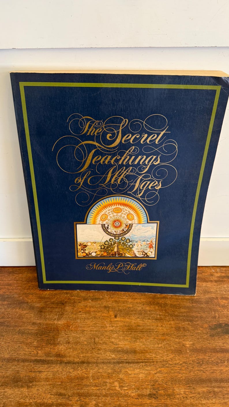 Może przedstawiać: Książka w twardej oprawie zatytułowana "The Secret Teachings of All Ages" autorstwa Manly'ego P. Halla. Książka ma ciemnoniebieską okładkę ze złotymi literami i kolorową ilustracją. Książka leży na drewnianej powierzchni.