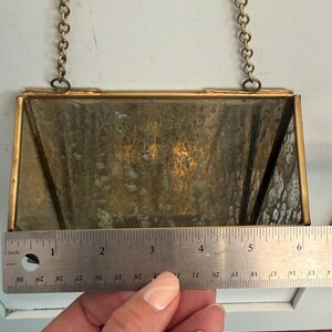 May include: Rectangular vintage brass-framed mirror with a chain for hanging. The mirror is approximately 5 inches wide. The glass has a mottled, aged appearance. A ruler is placed next to the mirror.