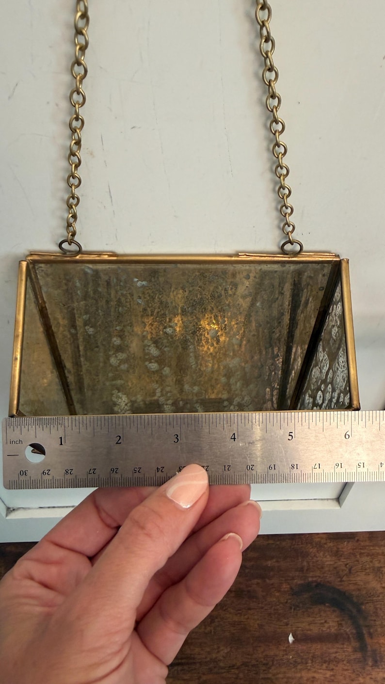 May include: Rectangular vintage brass-framed mirror with a chain for hanging. The mirror is approximately 5 inches wide. The glass has a mottled, aged appearance. A ruler is placed next to the mirror.