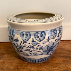 May include: A round, ceramic planter with a cream-colored rim and a blue and white floral design. The planter has a wide opening and a slightly curved body. The floral pattern includes flowers, leaves, and other decorative elements.