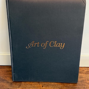 May include: A dark blue book titled "Art of Clay" in gold lettering. The book is rectangular and appears to be hardcover. The book is resting on a wooden surface.