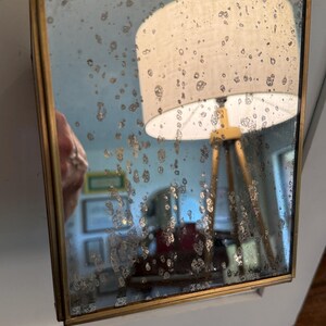 May include: Square antique mirror with a gold-colored metal frame, suspended by a delicate chain. The mirror's surface has a distressed, speckled appearance, reflecting a room with a lamp and other objects.