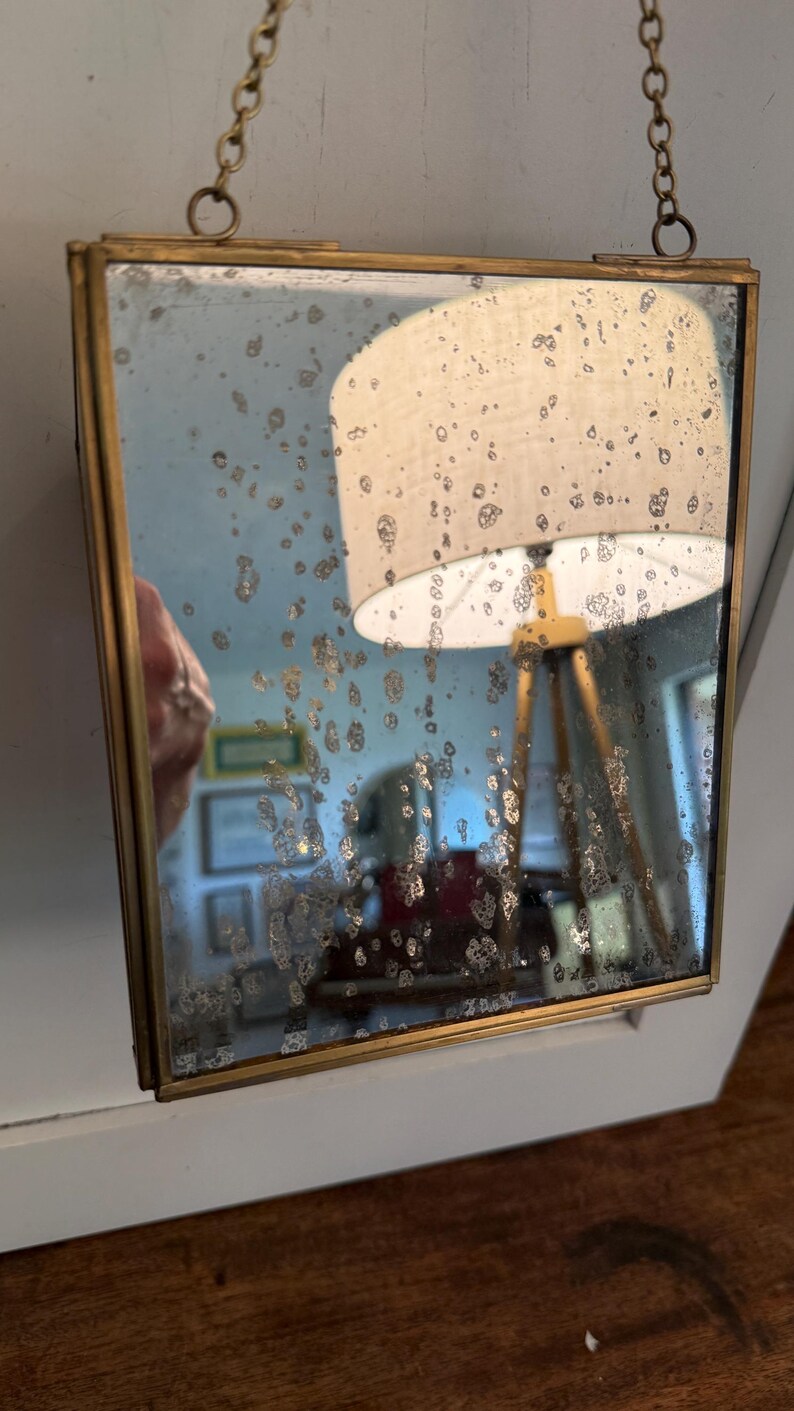 May include: Square antique mirror with a gold-colored metal frame, suspended by a delicate chain. The mirror's surface has a distressed, speckled appearance, reflecting a room with a lamp and other objects.