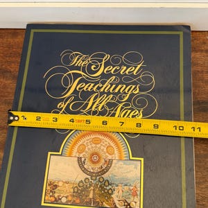 Może przedstawiać: Twarda oprawa książki zatytułowana "The Secret Teachings of All Ages" autorstwa Manly P. Halla. Książka jest ciemnoniebieska ze złotymi literami i kolorową ilustracją. Książka ma około 25 cm długości.