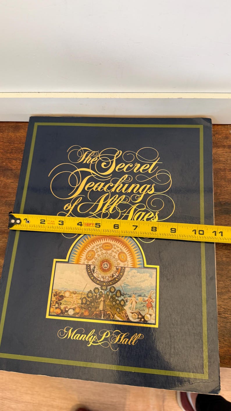 Może przedstawiać: Twarda oprawa książki zatytułowana "The Secret Teachings of All Ages" autorstwa Manly P. Halla. Książka jest ciemnoniebieska ze złotymi literami i kolorową ilustracją. Książka ma około 25 cm długości.