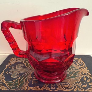 May include: A vibrant red glass pitcher with a handle and a spout. The pitcher has a faceted design and sits on a small base. The handle is also faceted. The pitcher is likely vintage and is a decorative item.