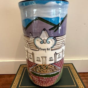 May include: A tall, hand-painted ceramic vase with a black rim. The vase features a landscape design with mountains, sky, and buildings. The text "Chimney Rock" is visible on the vase. The base of the vase has a red and green pattern.