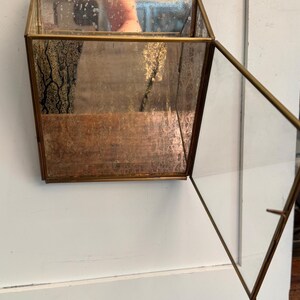 May include: A hanging, geometric glass and brass terrarium with a hinged door. The terrarium is suspended by a delicate chain. The glass has a mirrored effect, and the brass frame has a vintage appearance. The terrarium is empty.