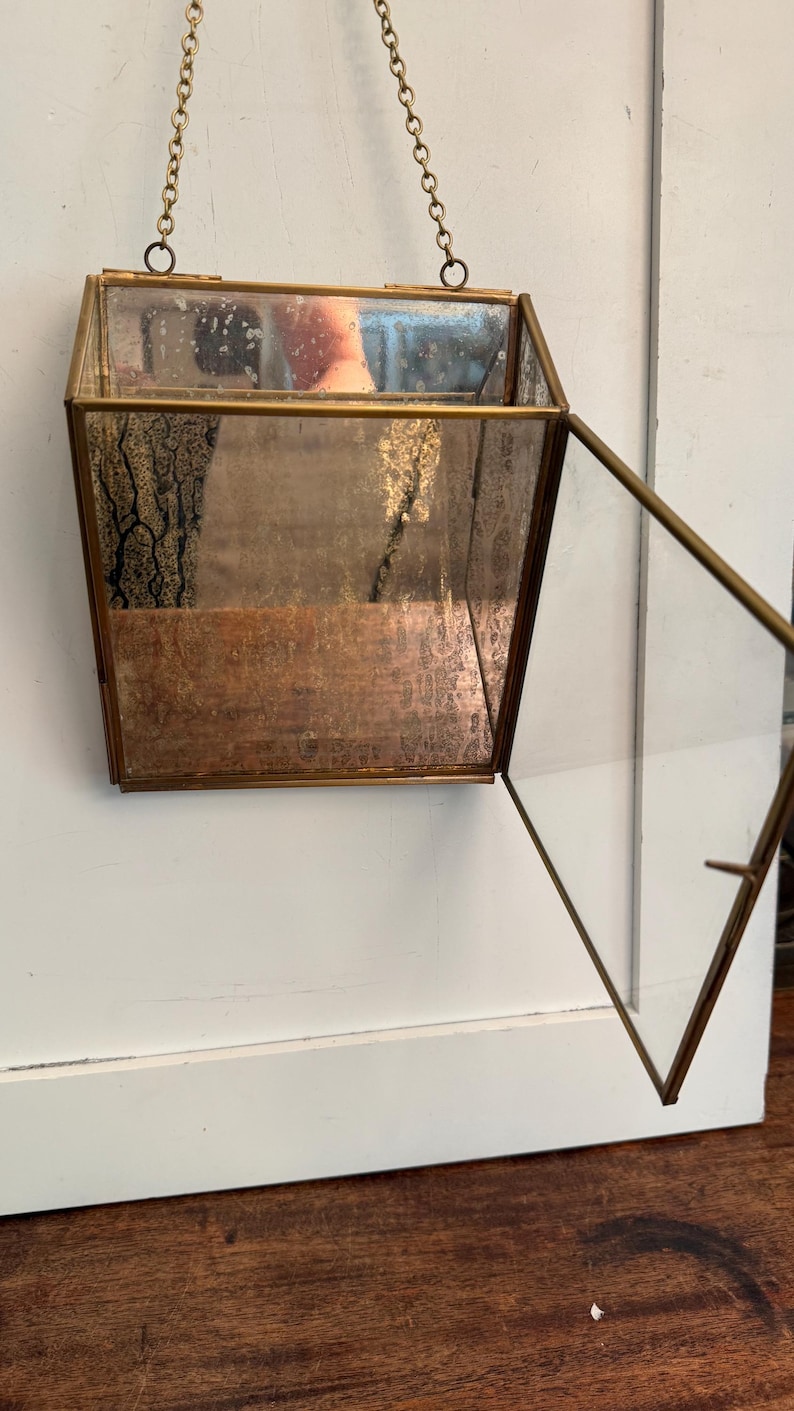 May include: A hanging, geometric glass and brass terrarium with a hinged door. The terrarium is suspended by a delicate chain. The glass has a mirrored effect, and the brass frame has a vintage appearance. The terrarium is empty.