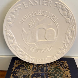 May include: A white ceramic Easter plate with embossed text and imagery. The plate features the words "EASTER", "JESUS IS NOT HERE", and "HE IS RISEN". A floral border surrounds a central scene of an empty tomb. The year "1972" is also present.