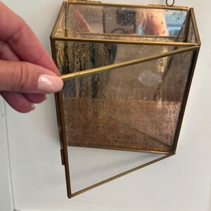 May include: A geometric, hanging wall planter with a brass frame and clear glass panels. The planter is suspended by a delicate gold chain. The interior of the planter has a textured, aged appearance. The open design allows for easy planting and display.