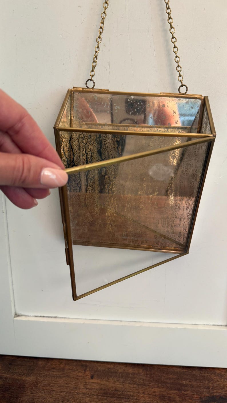 May include: A geometric, hanging wall planter with a brass frame and clear glass panels. The planter is suspended by a delicate gold chain. The interior of the planter has a textured, aged appearance. The open design allows for easy planting and display.