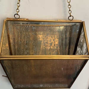 May include: A vintage hanging planter with a brass-colored frame and chain. The planter has a rectangular shape with angled sides and a tarnished, aged appearance. The glass panels are visible.