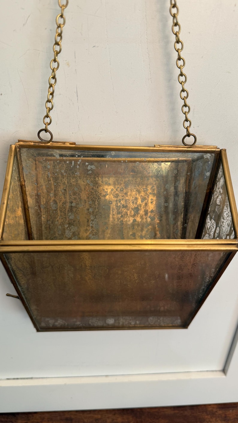 May include: A vintage hanging planter with a brass-colored frame and chain. The planter has a rectangular shape with angled sides and a tarnished, aged appearance. The glass panels are visible.
