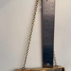 May include: A small, rectangular, hanging glass and brass terrarium. The terrarium is suspended by a brass chain and is approximately 4 inches in height. A metal ruler is placed next to the terrarium.