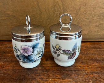 May include: Two white porcelain egg cups with silver-toned lids and handles. Each cup is decorated with a floral design in shades of purple, blue, and yellow. The egg cups are on a wooden surface.