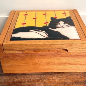 May include: A wooden box with a hinged lid. The lid features a ceramic tile with a black and white cat design on a yellow background with red accents. The box is made of light-colored wood.