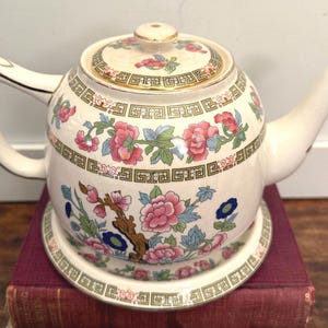 May include: Cream-colored ceramic teapot with a matching lid and saucer. The teapot features a floral design with pink and blue flowers, green leaves, and gold trim. The handle and spout are curved.