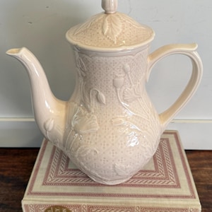 May include: A pale pink ceramic teapot with a matching lid and handle. The teapot features embossed floral and lattice patterns. It sits atop a book with a decorative cover. A classic piece for tea lovers.