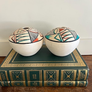 May include: Two spherical, off-white ceramic vases with geometric designs in black, teal, and orange. The vases sit atop a stack of green books with gold lettering and detailing. The vases have a small opening at the top.