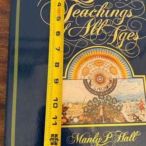 Może przedstawiać: Twarda oprawa książki zatytułowana "The Secret Teachings of All Ages" autorstwa Manly P. Hall. Książka jest ciemnoniebieska ze złotymi literami i szczeg&oacute;łową ilustracją. Książka ma około 35,5 cm wysokości.