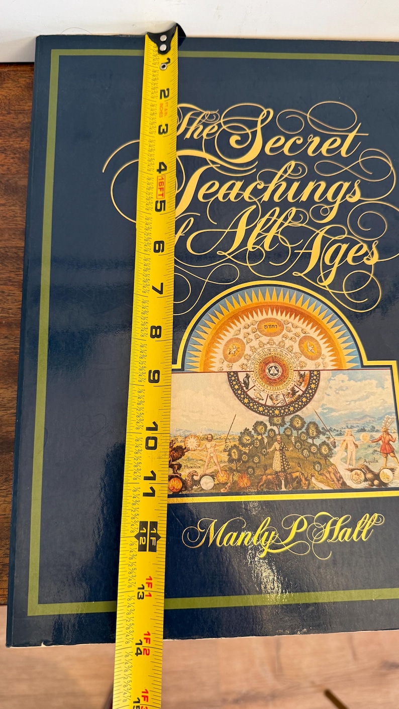 Może przedstawiać: Twarda oprawa książki zatytułowana "The Secret Teachings of All Ages" autorstwa Manly P. Hall. Książka jest ciemnoniebieska ze złotymi literami i szczeg&oacute;łową ilustracją. Książka ma około 35,5 cm wysokości.