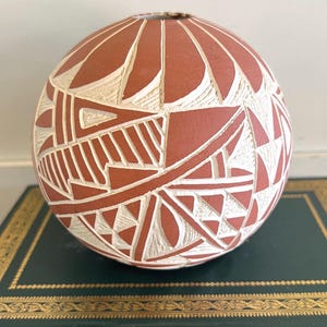 May include: A round, terracotta-colored ceramic vase with a carved geometric design. The design features white, angular shapes and lines, creating a contrasting pattern across the surface. The vase is displayed on a dark green surface.