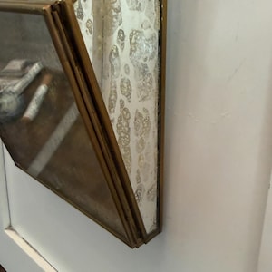 May include: Geometric hanging terrarium with a brass frame and distressed glass panels. The terrarium is suspended by a chain and is mounted on a white wall. The glass has a unique, aged appearance.