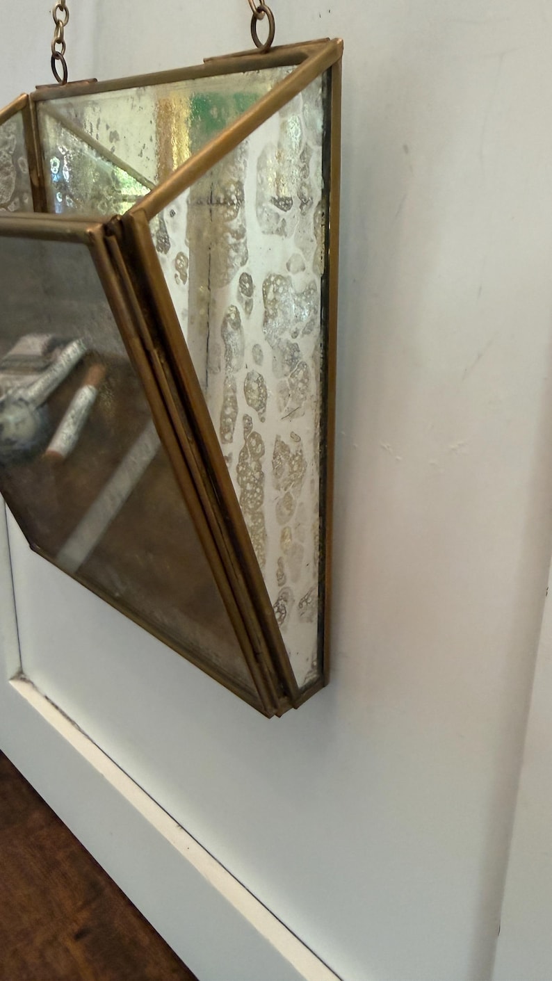 May include: Geometric hanging terrarium with a brass frame and distressed glass panels. The terrarium is suspended by a chain and is mounted on a white wall. The glass has a unique, aged appearance.