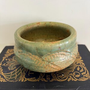 May include: A small, round ceramic bowl with a textured, leaf-like design around the exterior. The bowl has a green and brown color scheme, with a speckled appearance. The interior is a darker green. The bowl is sitting on a dark blue book.