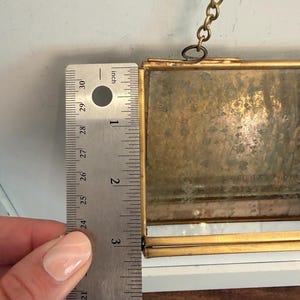 May include: A rectangular, antique-style picture frame with a gold-colored metal frame. The frame is approximately 5 inches (12.7 cm) tall. A metal ruler is placed next to the frame for measurement. A gold chain is attached to the top.