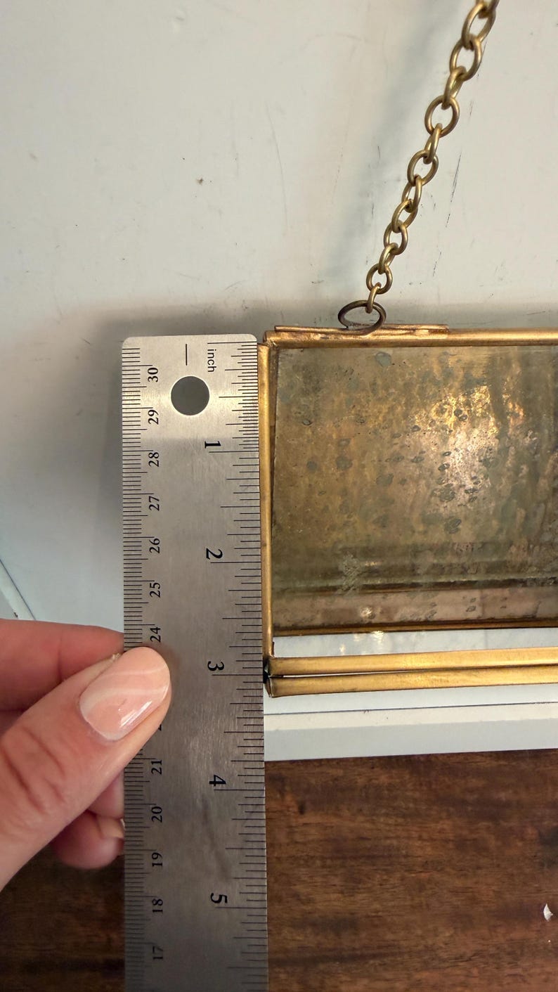 May include: A rectangular, antique-style picture frame with a gold-colored metal frame. The frame is approximately 5 inches (12.7 cm) tall. A metal ruler is placed next to the frame for measurement. A gold chain is attached to the top.