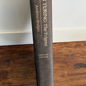 May include: A hardcover book titled "ALAN TURING: The Enigma" by Andrew Hodges. The book's spine is dark gray with gold lettering and the publisher's name, Simon and Schuster, at the bottom. The book is held against a white wall and a wooden surface.