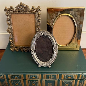 May include: Three antique picture frames in gold and silver tones. The frames are rectangular and oval, with ornate detailing. The frames are displayed on top of two green books with gold lettering.