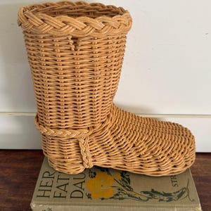 May include: A woven wicker boot-shaped basket, light brown in color, with a braided rim. The basket is sitting on top of a vintage book with the title "Her Father's Daughter". The boot is approximately 10 inches tall.