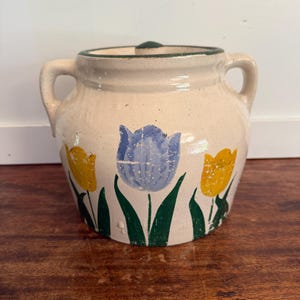 May include: Cream-colored ceramic jar with a lid and two handles. The jar is decorated with yellow and blue tulip flower designs and green leaves. The rim and lid edge are trimmed with green.