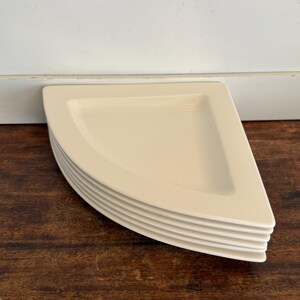 May include: A stack of six off-white, quarter-circle shaped plates. The plates are stacked on a dark brown wooden surface, with a white wall and trim in the background. The plates have a slightly raised edge.