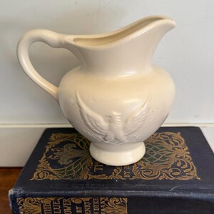 May include: Cream-colored ceramic pitcher with a curved handle and a spout. The pitcher features an embossed design of a bird with outstretched wings on the front. It is sitting on top of a stack of antique books with gold lettering.