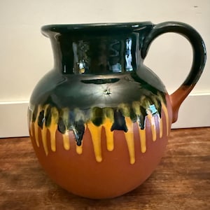 May include: A ceramic pitcher with a dark green glazed top and handle. The body is a warm brown with yellow and dark green drips. The pitcher has a rounded shape and a large handle.