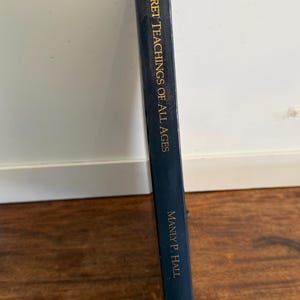 Może przedstawiać: Ciemnoniebieska książka zatytułowana "Secret Teachings of All Ages" autorstwa Manly P. Halla. Tytuł książki i nazwisko autora są w złotych literach. Książka jest trzymana pionowo.